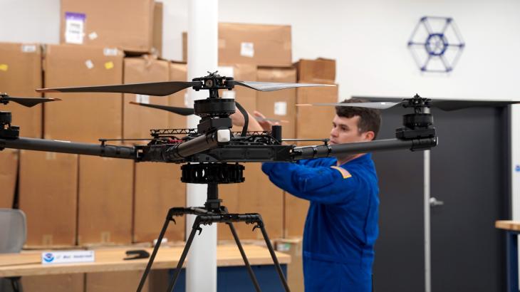 A NOAA corps officer in a blue flight suite adjusts the blades of an uncrewed aircraft on a work bench